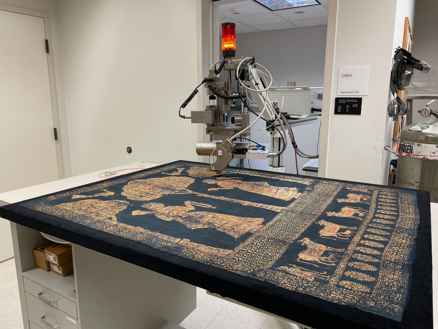 The arm of a machine reaches out over a blue-black and gold painting spread across a table in a white room.