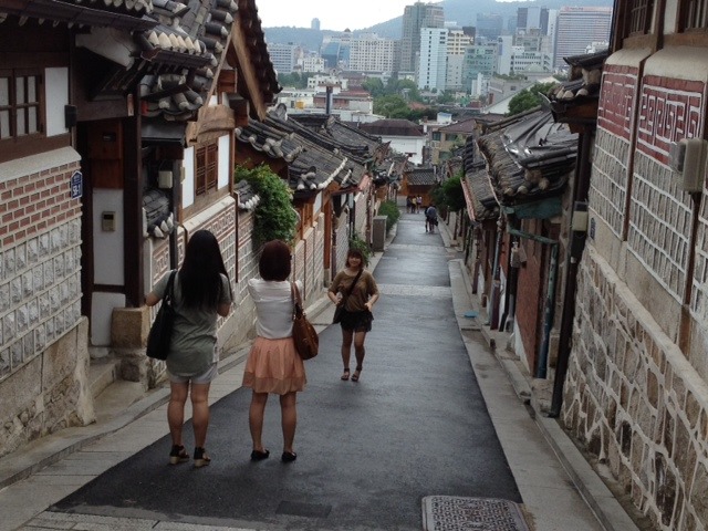 Street with rows of brick buildings on both sides and city buildings in the distance.