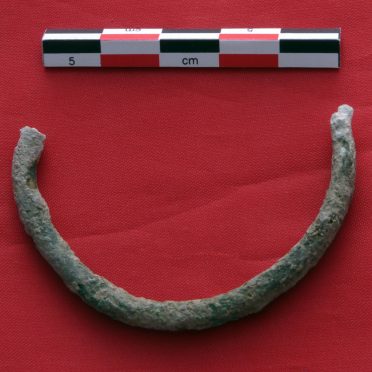 Half of a broken bronze bracelet, covered in green and white patina, next to an archaeological centimeter scale, on a red surface.