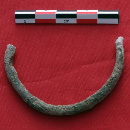 Half of a broken bronze bracelet, covered in green and white patina, next to an archaeological centimeter scale, on a red surface.