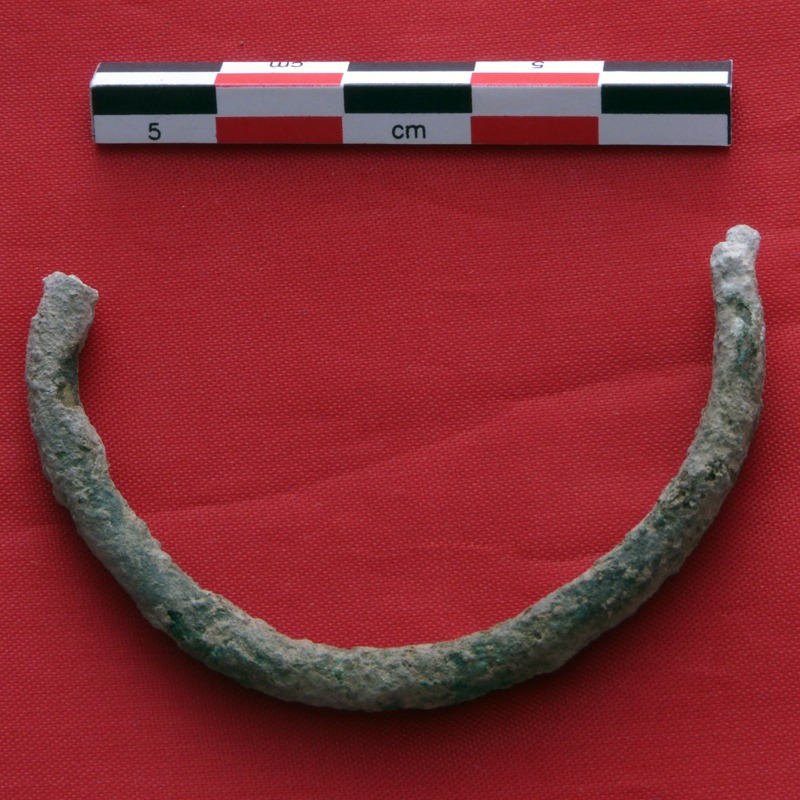 Half of a broken bronze bracelet, covered in green and white patina, next to an archaeological centimeter scale, on a red surface.
