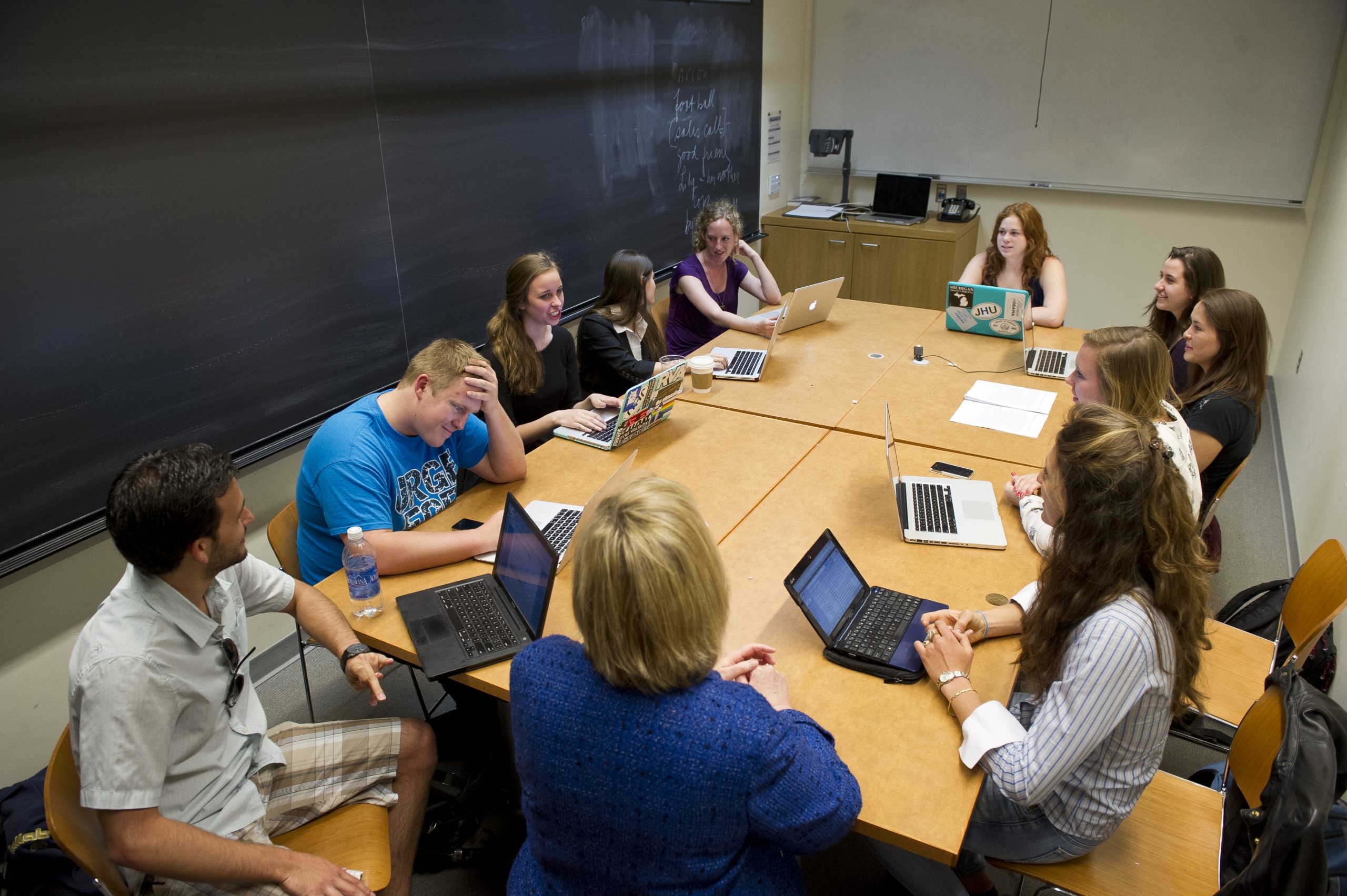 A group of people in a classroom sat around a wooden table with laptops in front of them.
