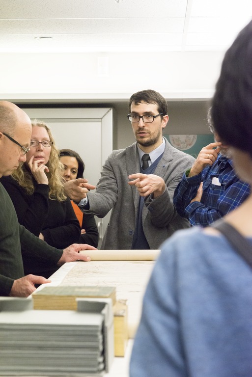 A man in a suit holding his hands out over a scroll towards an small group of onlookers.