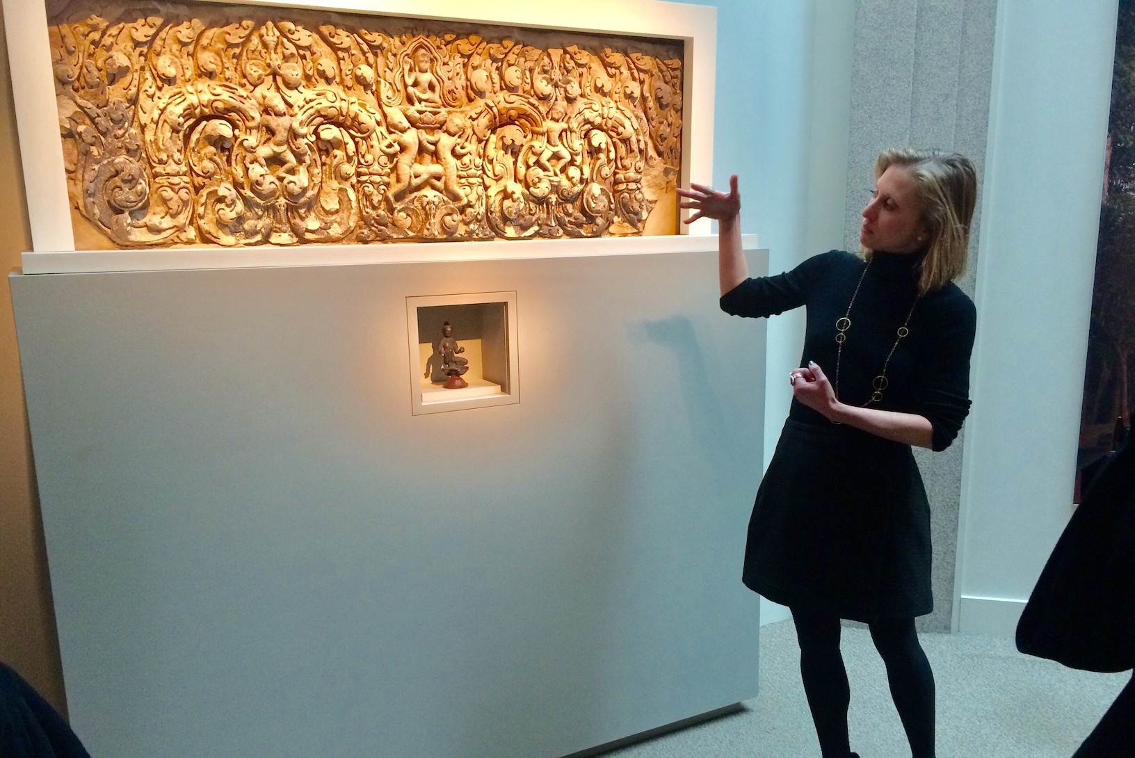 A woman gestures toward a stone tablet hanging on the wall.