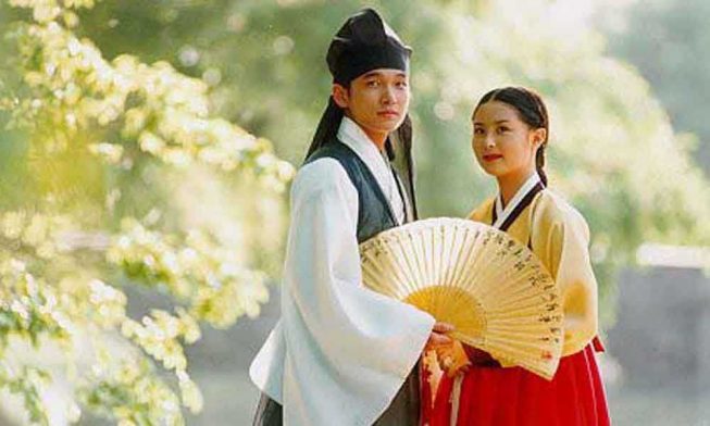 A man and woman standing close together looking into the distance in the foreground. The man holds an open fan at the waist.