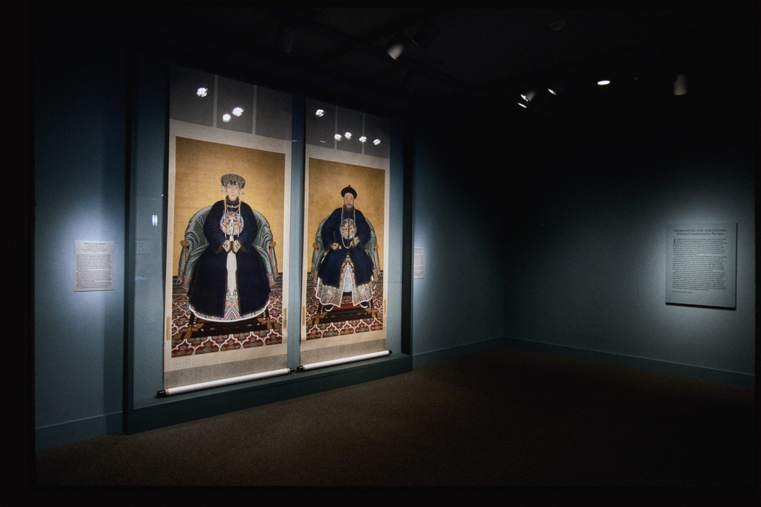 Two large Chinese ancestor portraits hang on a gallery wall in a starkly lit room painted blue.