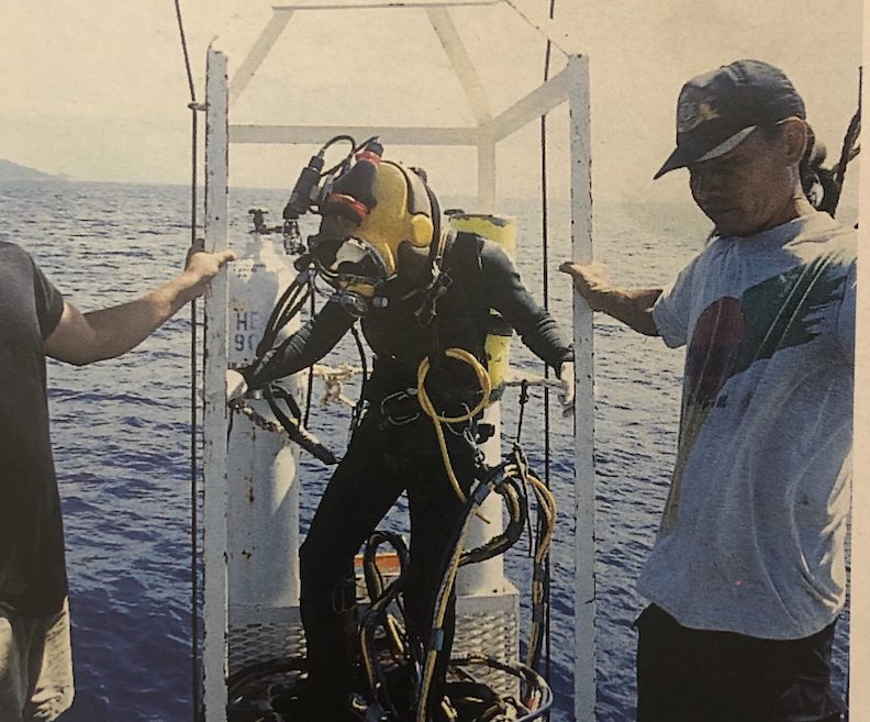 A man wearing a black wetsuit and yellow helmet and tank preparing to dive into sea water. 