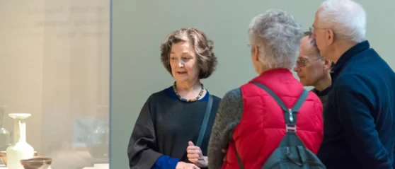 A woman in a black long sleeve shirt stands near a white ceramic vase in a glass case, she faces a group of three people and is speaking to them about the object in the case.