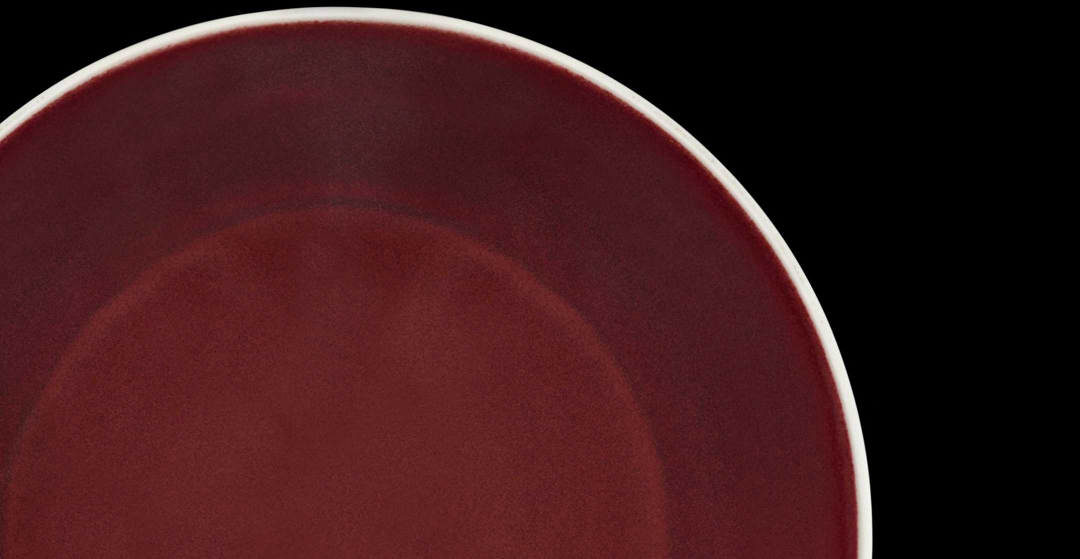 A partial view of a ceramic dish with a deep red glaze and crisp, white border against a black background.