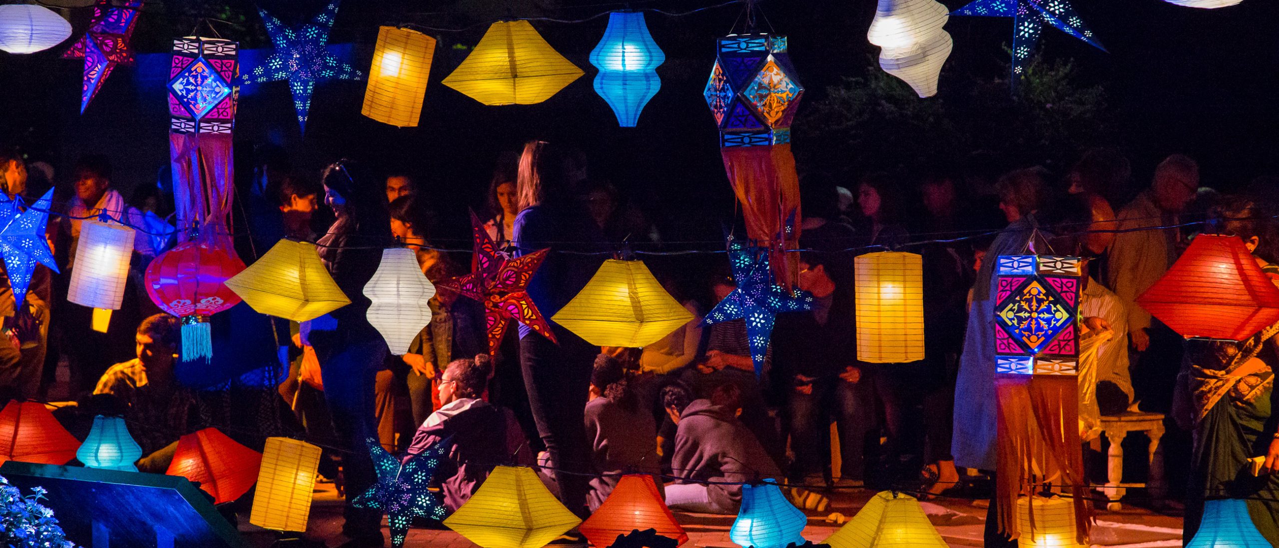 Colorful lit-up lanterns hanging outside with a crowd of people gathered in the background.