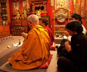 four People, led by a man in the orange robes of a Buddhist monk, kneel with their hands tented in prayer in an opulent red and gold room filled with a gong, sculptures, hanging scrolls, and bells.