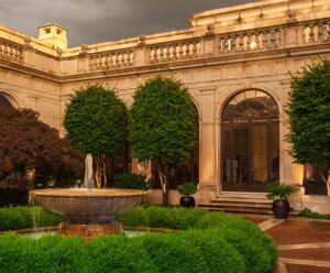 A brick-paved and tree-lined courtyard of a neoclassical building, illuminated and seen against a dusky sky