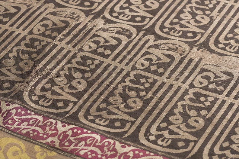 A dark textile, viewed at an angle, with lighter Arabic text offset, and a purple border also with white inscriptions.