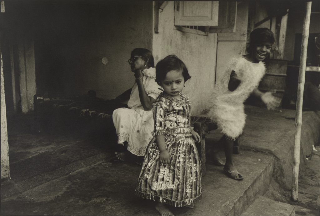 A young child appears finely dressed on a doorstep and presents a contrast to the two figures behind her. While her youthful countenance sets her apart from the bored woman on the left, her still and thoughtful demeanor is a foil to the smiling girl in an excitable blur of motion on the right.