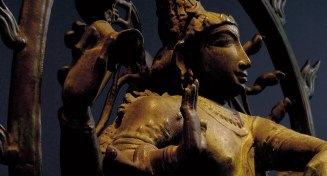 A close-up view of a bronze sculpture, lit with warm light and viewed in three-quarter view, of a figure with a raised hand, against a dark blue background.