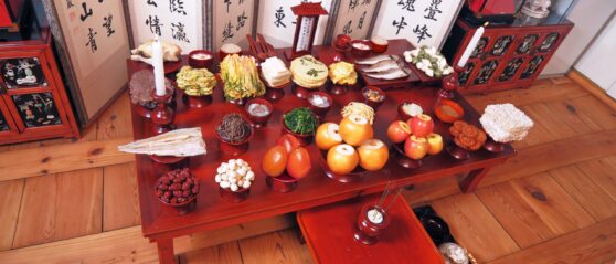 a red table filled with food, candles, incense. a screen with writing stands behind it