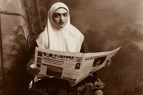 Seated woman, wearing a white head covering, holding open a newspaper