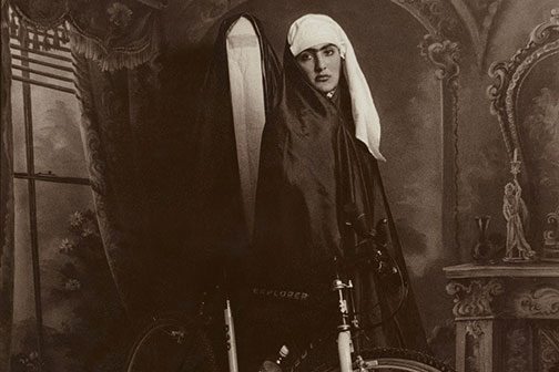 Black and white photo of a woman wearing a white head covering and black cloak, standing next to a bicycle, in front of a painted faux interior background