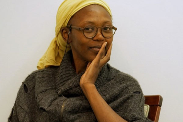 woman in a yellow headscarf, glasses and green top touches her cheek and smiles at the viewer.