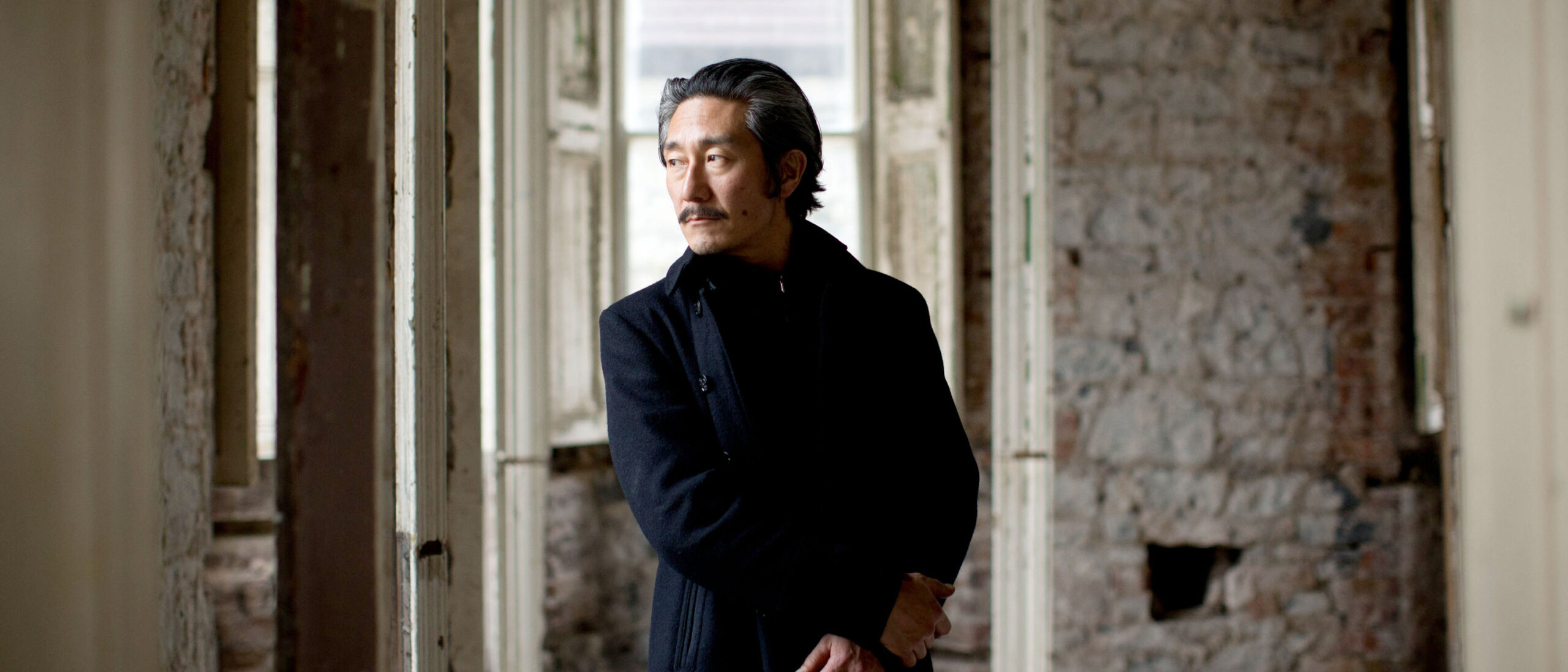 artist michael joo, a man with a mustache in a navy coat, stands in an old building with peeling ivory paint.