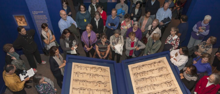 A top-down view of a crowd of people gathered around and looking at two very large folios of manuscript calligraphy.