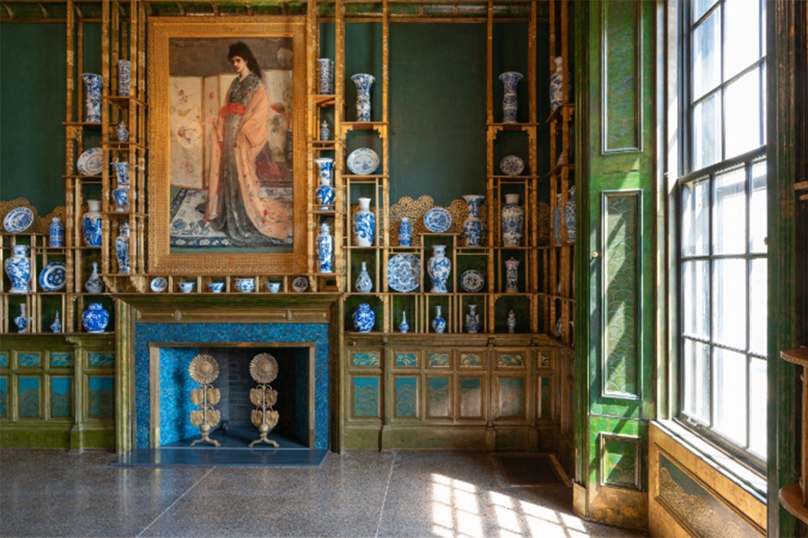 Empty room with ornate decorated shelves displaying a number of blue-and-white ceramic vessels with a painting of a woman above a fireplace. On the right, a window with open shutters lets natural light into the room.