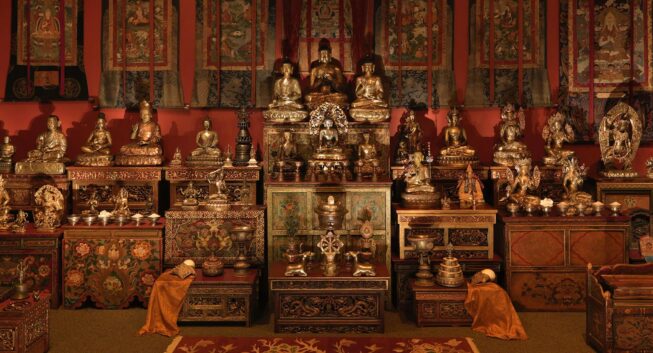 A dense arrangement of golden figural statues, vessels, altars, and textile wall hangings, displayed in a warmly lit, red-colored room.