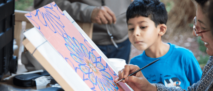 A smiling artist works on a painted design as a child looks on