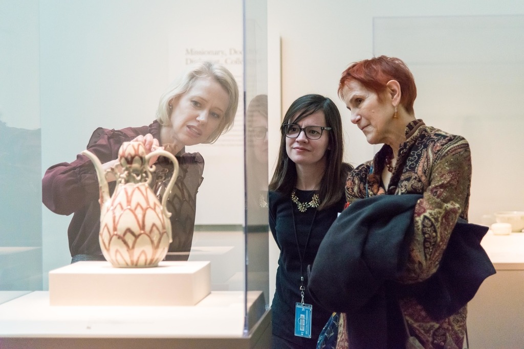 A woman with blonde hair points at a ceramic in a display case. Two woman standing on her right observe.