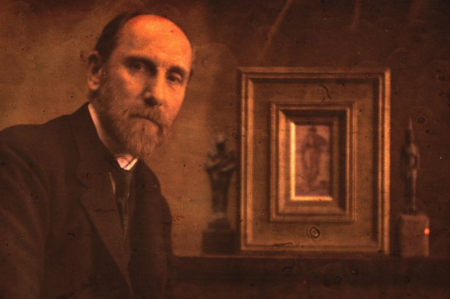 a bearded man in a suit leans next to two sculptures and a painting in an ornate gold frame.