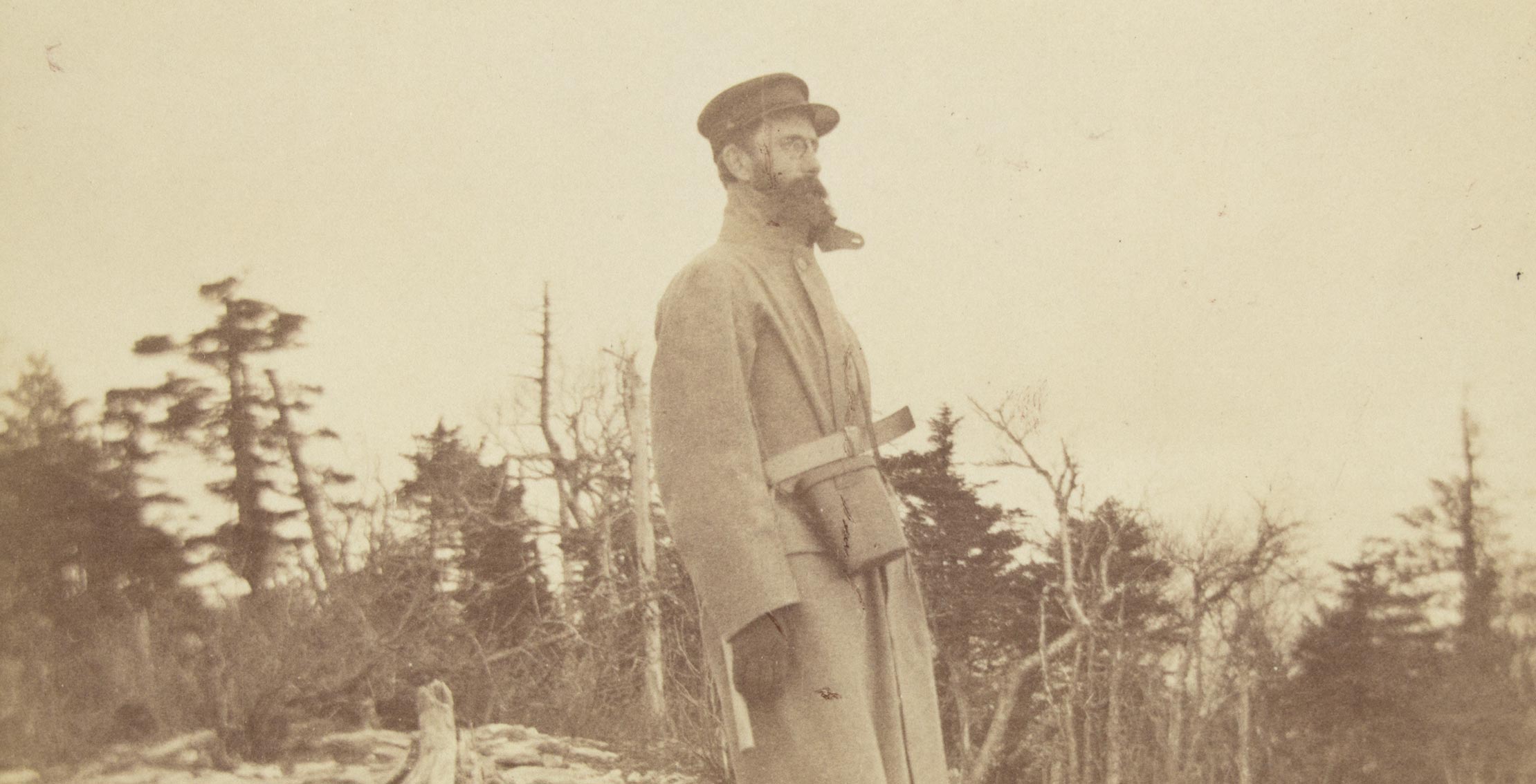 Sepia toned image of a man seen in profile, wearing a cap and long coat, trees along the horizon behind him.