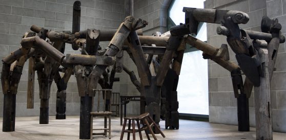 Tall wooden parts connecting and arranged in various angles in a spacious room. Wooden table and chairs stand at various points of the base of the larger wooden object.