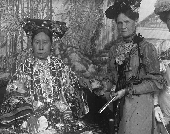 A photograph of two woman, the one on the left sits while the other stands by her side on the right. Their hands are joined.