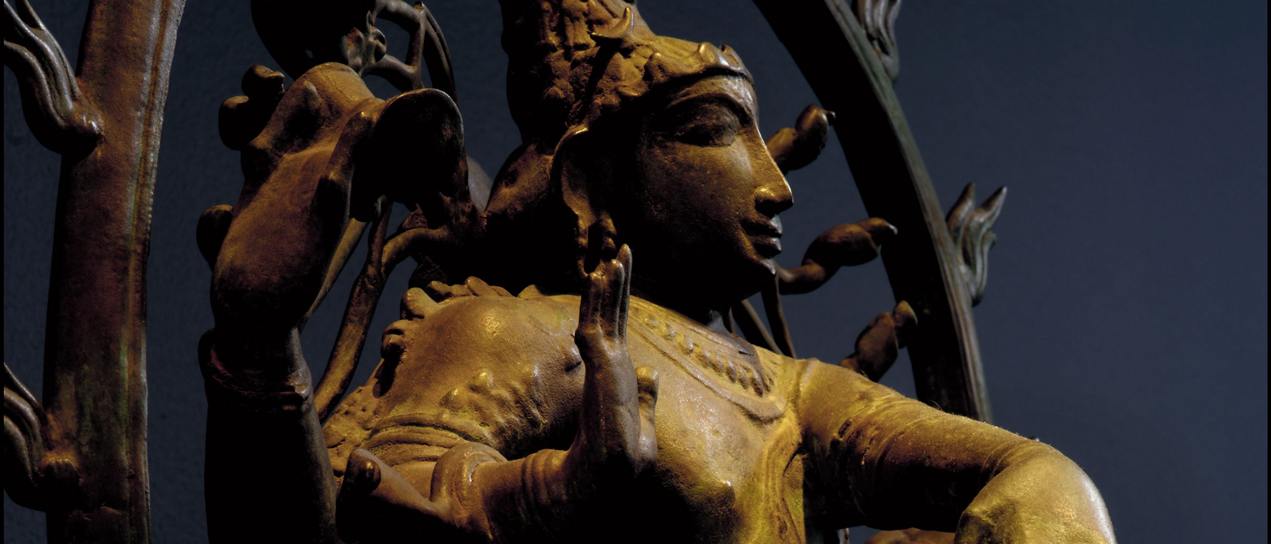 A close-up view of a bronze sculpture, lit with warm light and in three-quarter view, of a figure with a raised hand against a dark blue background.