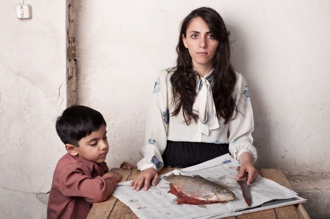 a dead fish on newspaper, a woman with a knife, and a young boy looking at the dead fish