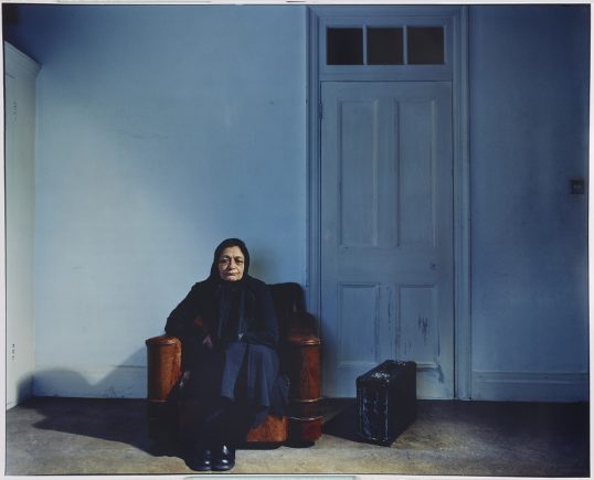 Elderly woman seated in an arm chair, with a suitcase, next to a door