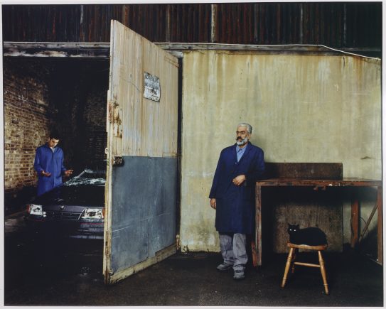 Ma leaning on a desk next to a garage door, which is partly open, revealing a car and a man in a blue mechanics jumpsuit beyond