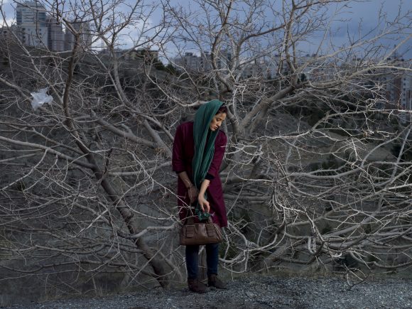 woman in a teal headscarf and burgundy outfit clutching a purse, surrounded by grey, naked trees, almost closing in on her.