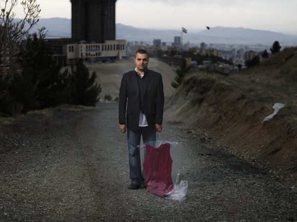 Man standing in the middle of a road, holding a clear garment bag containing a burgundy dress