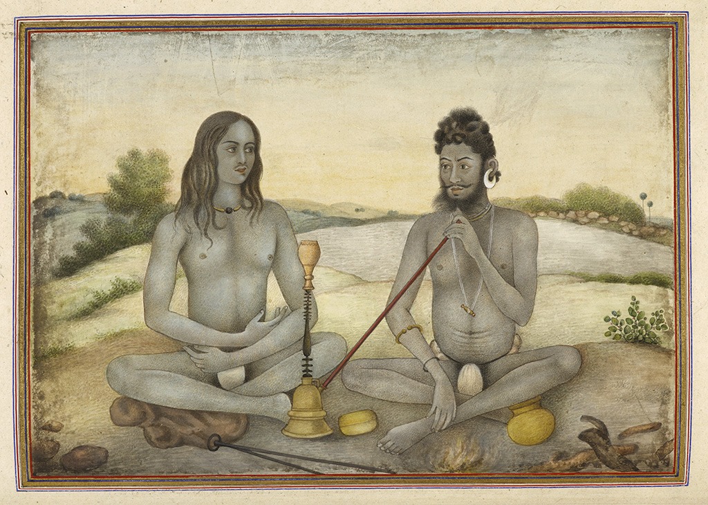 two men sitting cross-legged, the man wearing earrings smokes a pipe