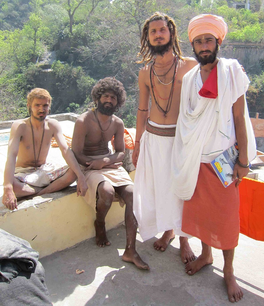 a group of bearded men, two standing, one sitting on a ledge and one sitting cross-legged on a ledge