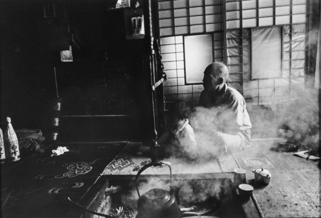 black and white photo of a man and a small child sitting near a steaming pot on the hearth, a tea set sitting ready near them