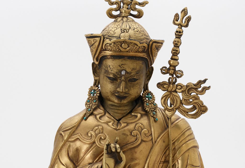 Gilt bronze sculpture of a meditating figure holding its hand up in a mudra. It has a pearl inset on its forehead, and turquoise earrings.