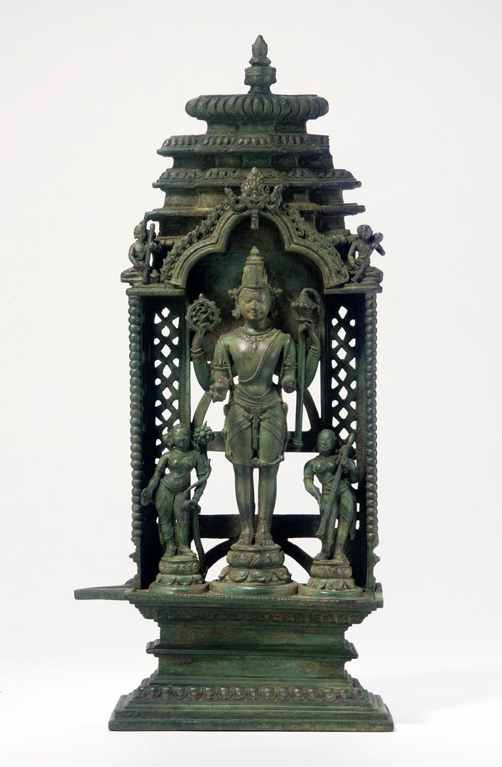 A bronze sculpture with a tall figure standing within a decorate structure, smaller female figures on either side of him.