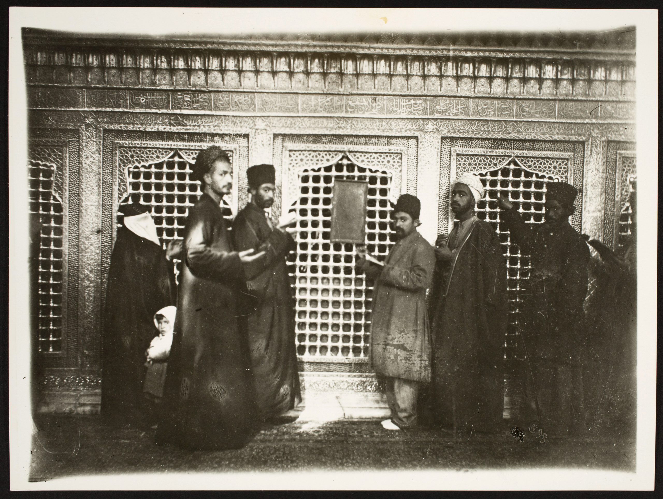 A group of pilgrims in dark clothes stand in front of the grated lattice of a shrine, looking at the camera.