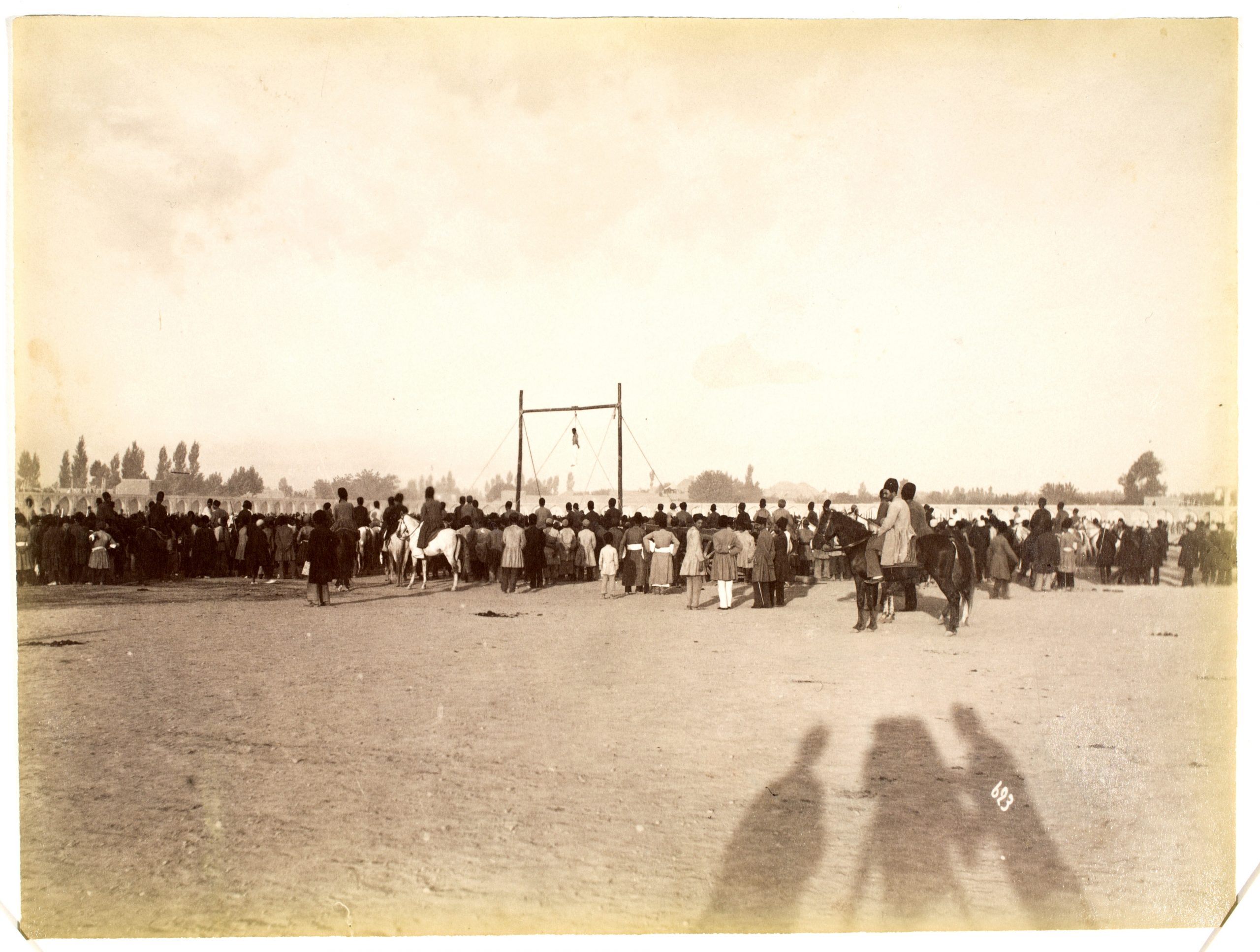 A figure hangs from gallows in the distance, surrounded by a large crowd of onlookers. A shadow of the photographer, the camera, and another figure are cast in the foreground.