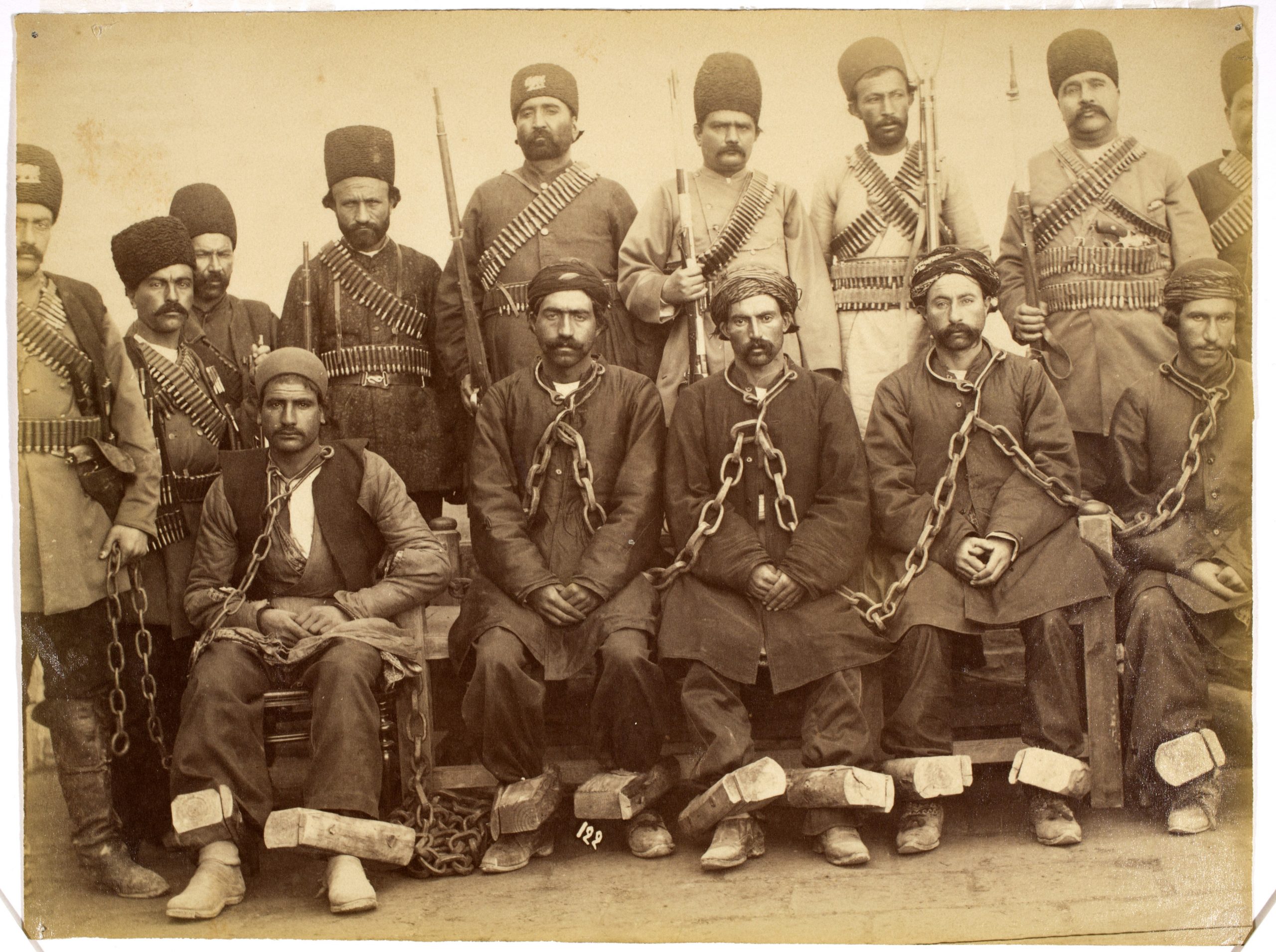 A line of prisoners with thick chains around their neck and blocks around their ankles sit in front of a row of standing guards who all hold muskets and wear bandoliers of bullets.