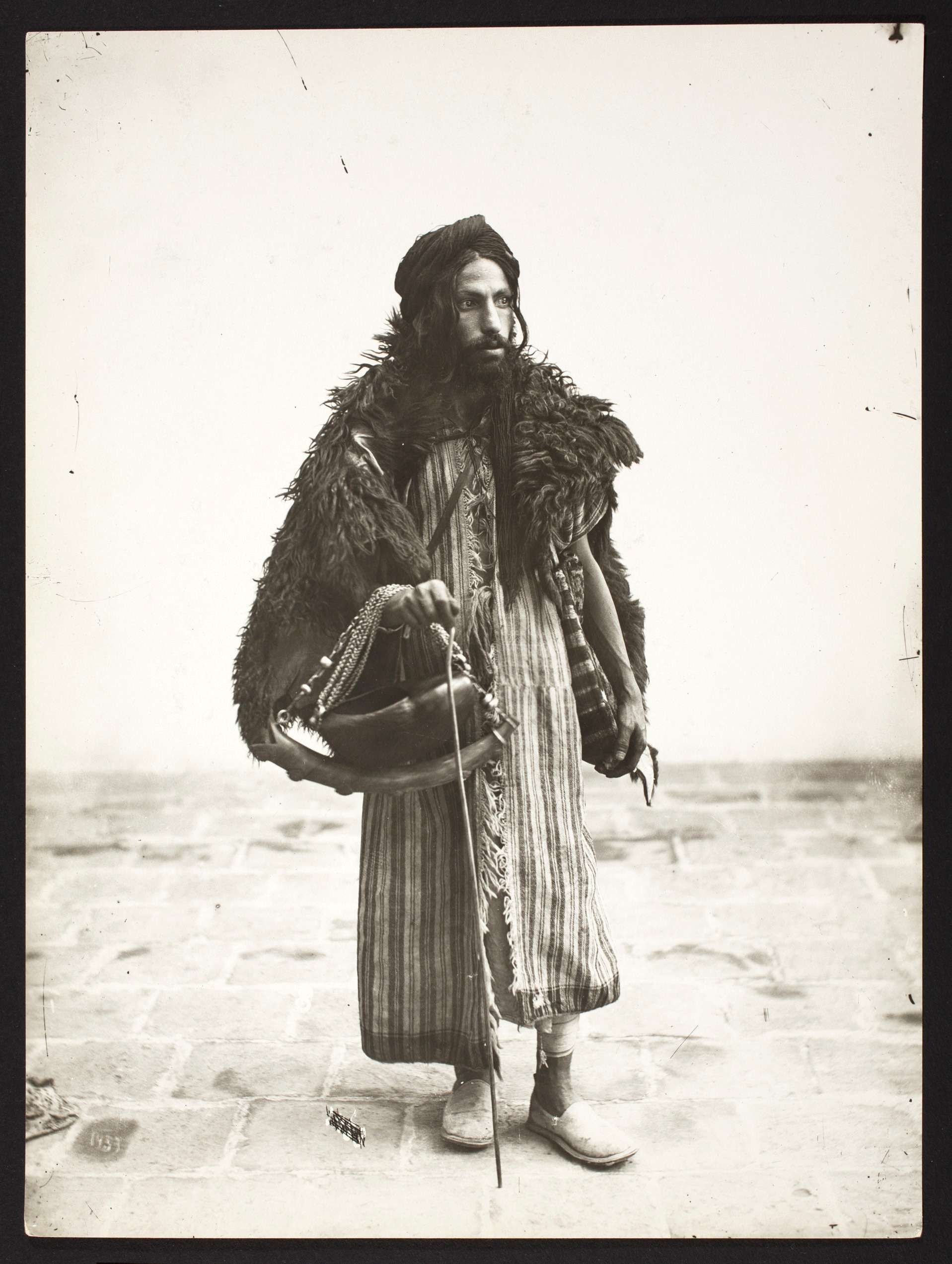 A man stands tensely, gaze avoiding the camera. Tresses of dark hair fall out from the front of his turban. He wears a vertically striped garment with a rough fur shawl. One hand holds a slim, hooked cane. A bowl hangs from a chain strap over the same arm.