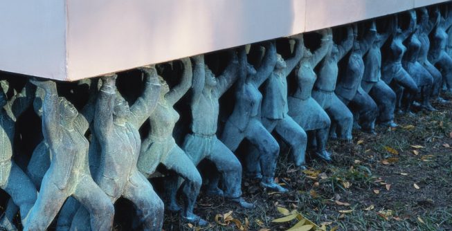 A row of sculpted figures, green with patina, all side by side in the grass and posed in active stances as they stretch their arms up to support a large white stone slab.