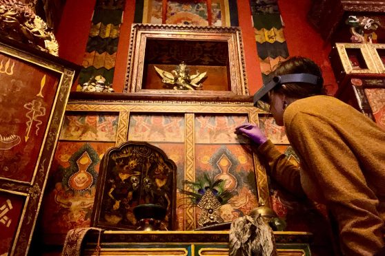 A scientist wearing an orange sweater and an optivisor bends close to a painted wall panel in an intricately decorated red-and-gold room.